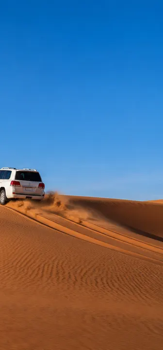 A white 4x4 driving over a red sand dune in the Dubai desert under a blue sky